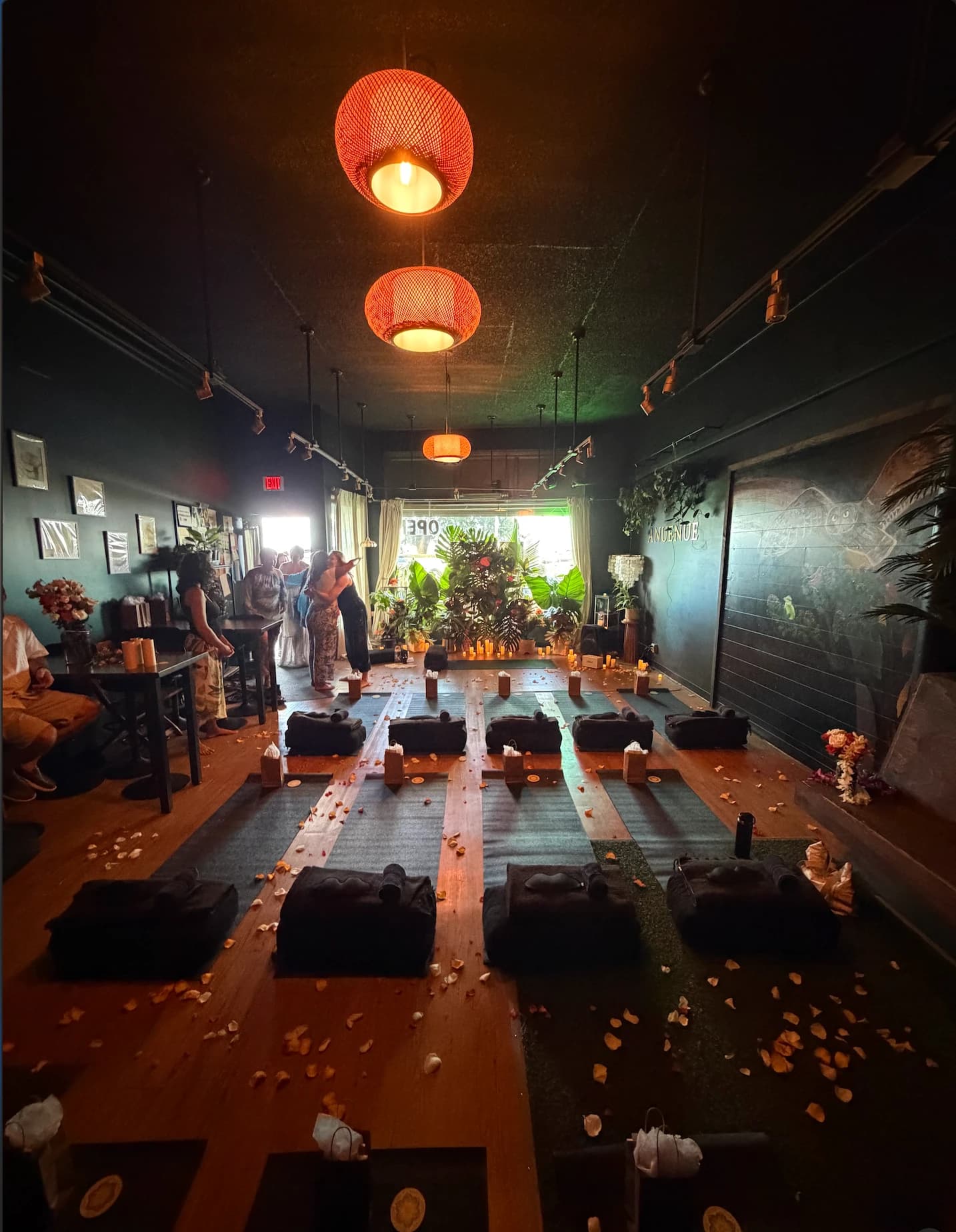 The lounge set for a candlelit sound bath, with yoga mats and bolsters on the wood floor, flower petals, amber lanterns overhead, and the owl mural on the wall