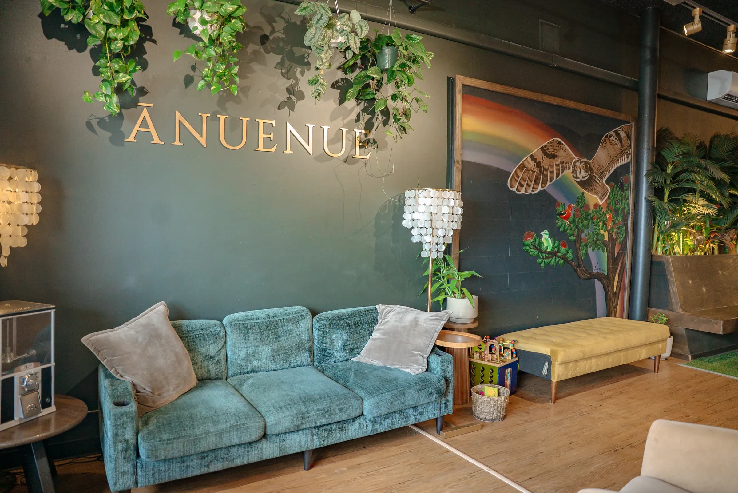 The lounge at Ānuenue On The Bay: brass lettering and trailing plants above a teal velvet couch, beside a painted rainbow and pueo mural